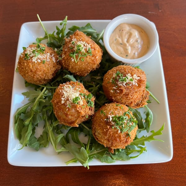 Five golden-brown arancini balls on a bed of arugula with a side of dipping sauce.