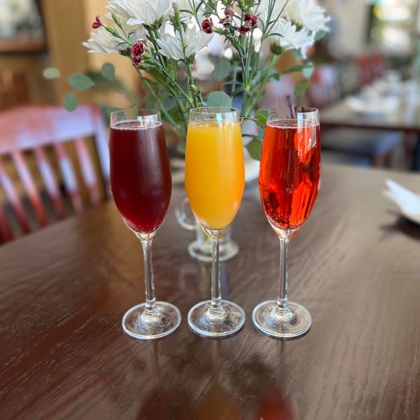 Three colorful mimosas, red, orange, and pink, on a wooden table with a floral centerpiece.