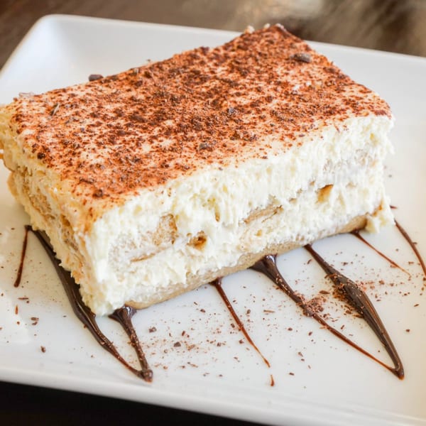 A rectangular slice of tiramisu with cocoa powder and chocolate drizzle on a white plate.