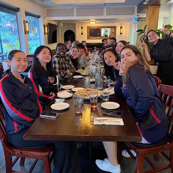 A large group of young people and staff enjoying pizza at a long wooden table inside Red Rooster Brick Oven.