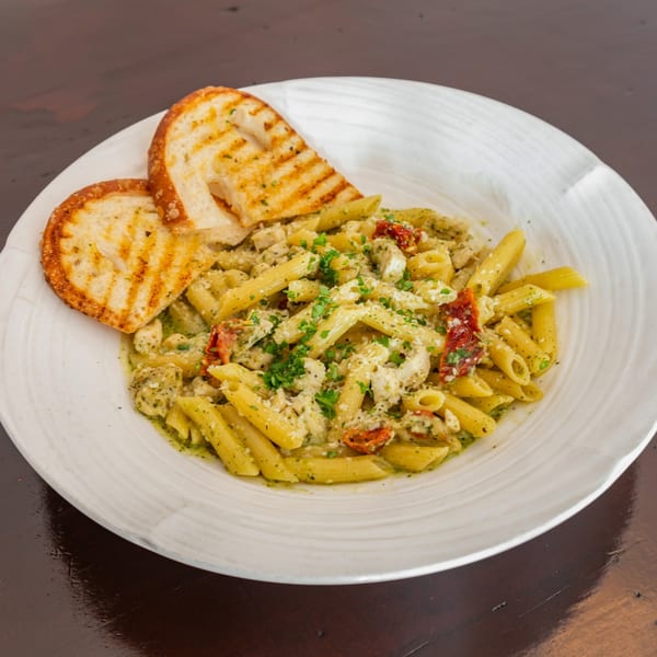 Penne pasta with pesto, chicken, sun-dried tomatoes, and garlic bread on a white plate.